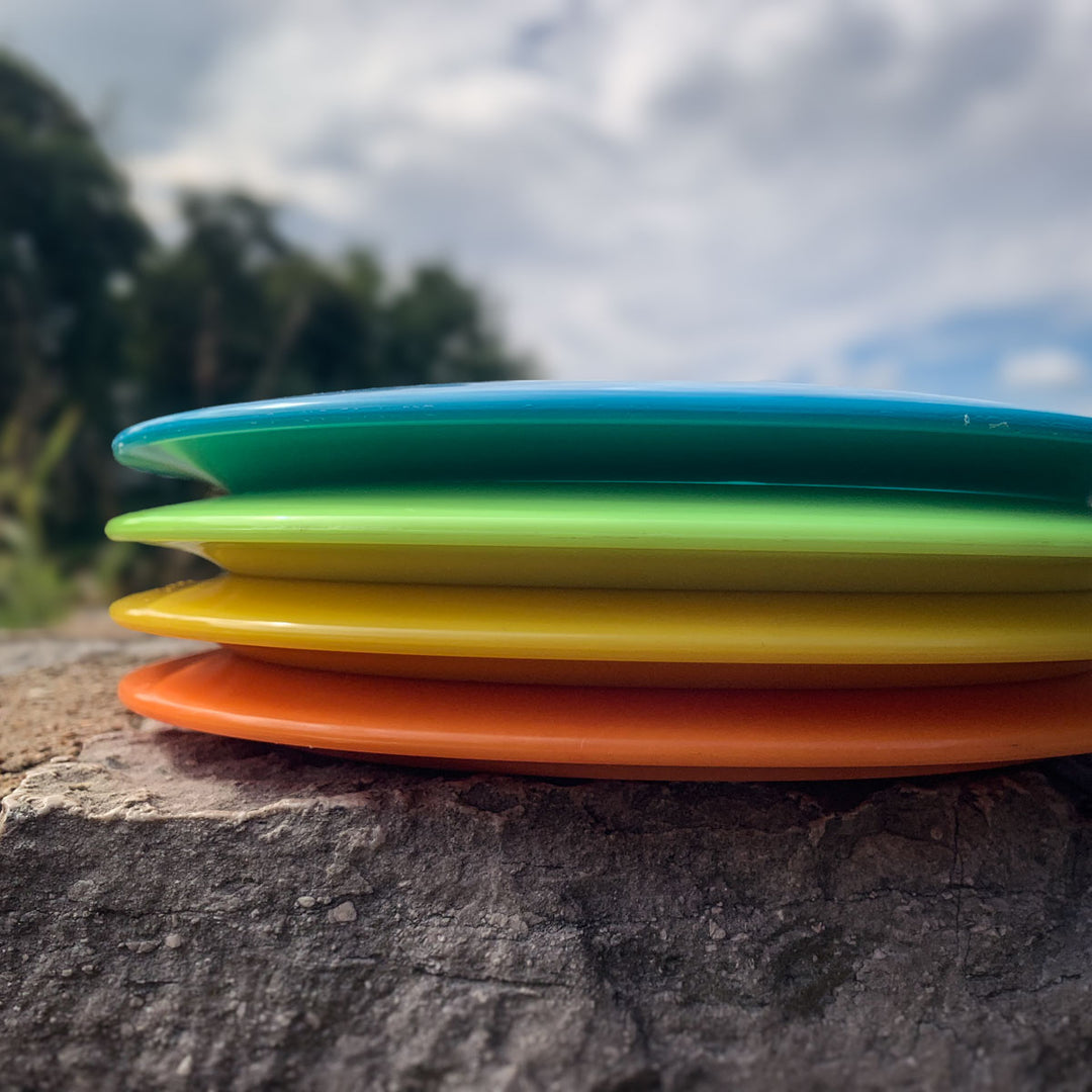 Four colorful surfboards—blue, green, yellow, and orange—stacked on rocks outdoors with the blurred silhouette of a Prototype 136 - NXT (Javelin) in the background among trees and sky.