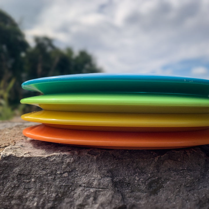 Four colorful surfboards—blue, green, yellow, and orange—stacked on rocks outdoors with the blurred silhouette of a Prototype 136 - NXT (Javelin) in the background among trees and sky.