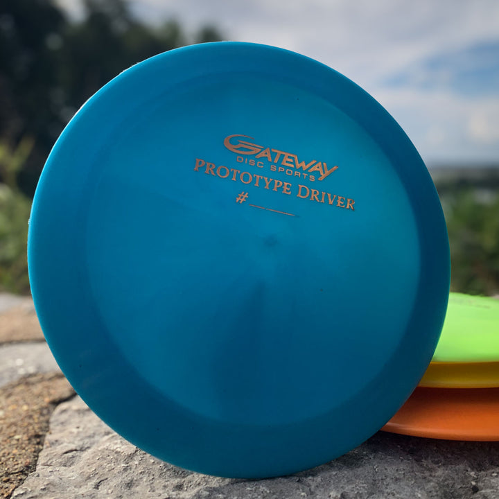 A blue Prototype 136 - NXT (Javelin) distance control driver rests on a rock, with other discs partially visible behind it.