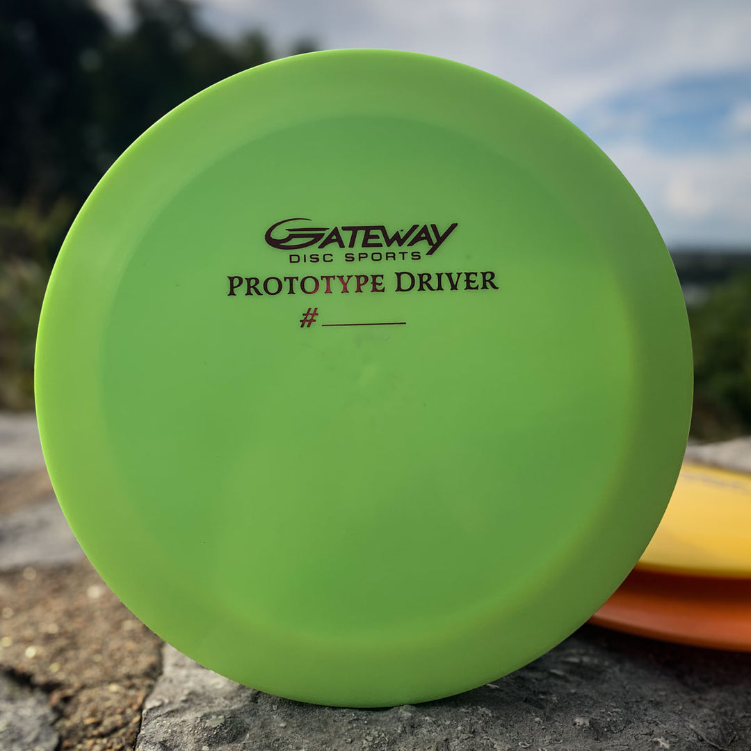 A green Prototype 136 - NXT (Javelin) disc is displayed upright outdoors on a rock—ideal as a distance control driver or for players wanting an aggressive driver on the course.