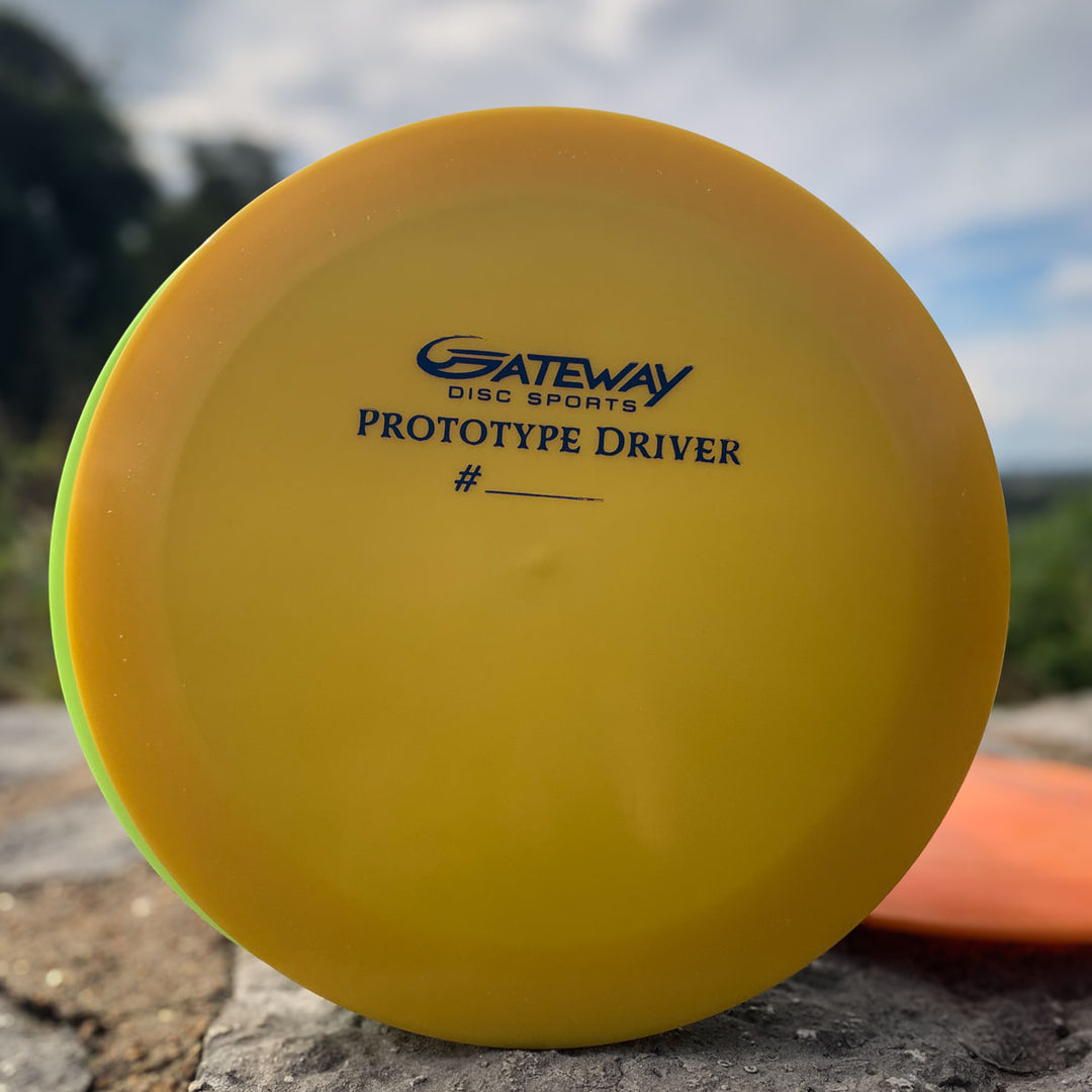 A yellow Prototype 136 - NXT (Javelin) disc golf driver labeled —Gateway Disc Sports Prototype Driver #____—� rests on a stone surface outdoors, poised to demonstrate its distance control and aggressive driver performance.