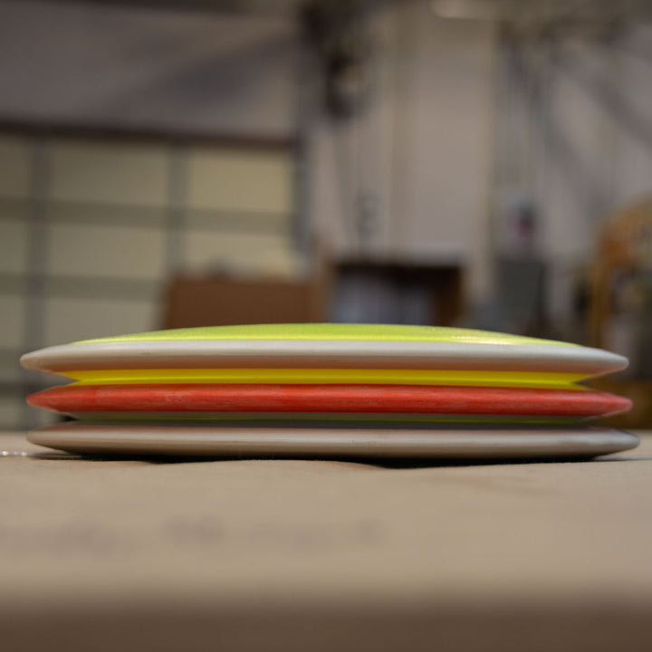 Three Prototype 93 surfboards with neon yellow, orange, and white rails are stacked horizontally on a flat surface, highlighting expert craftsmanship against the blurred background of a workshop.