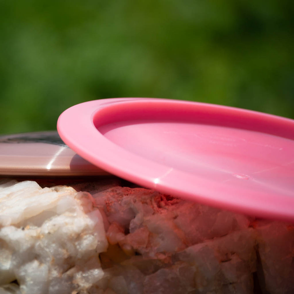 A close-up of a pink Reptilian DG Chameleon — Armor fairway driver disc lying on a rock, with greenery blurred in the background.