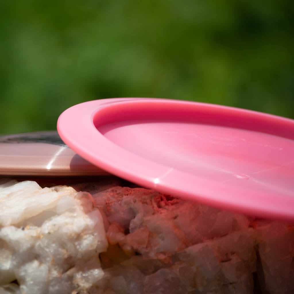 A close-up of a pink Reptilian DG Chameleon — Armor fairway driver disc lying on a rock, with greenery blurred in the background.