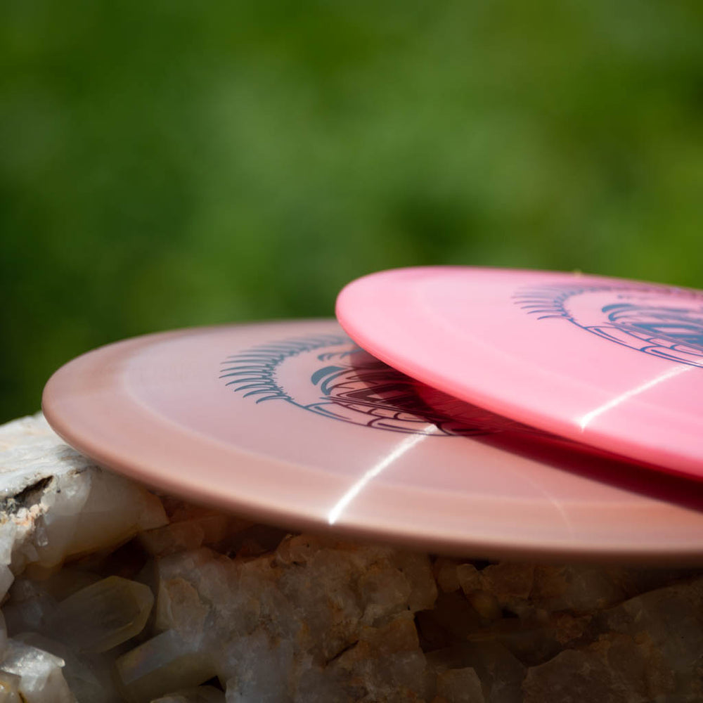 Two Reptilian DG Chameleon — Armor discs, one pink and one light brown fairway driver, sit on a rock with a blurred green background—ideal for mastering turn and fade.