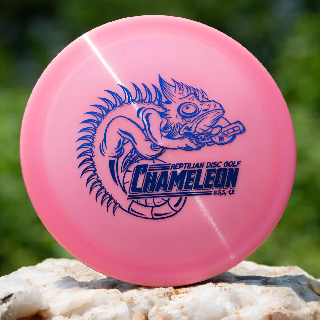A pink Reptilian DG Chameleon — Armor fairway driver disc golf frisbee with a blue chameleon illustration and —Reptilian Disc Golf Chameleon—� text printed on it rests on a rock outdoors. Ideal for players wanting balanced turn and fade.