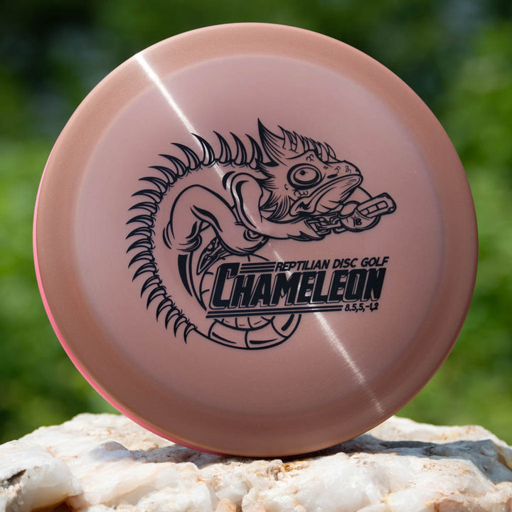 A pink Reptilian DG Chameleon — Armor fairway driver with a black chameleon illustration and "Reptilian Disc Golf Chameleon 8.5-5-1-2" text, emphasizing its turn and fade, shown on a rock background.