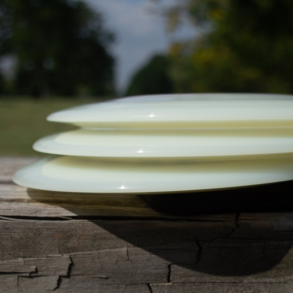 Three Realm - NXT Superglow distance drivers from Gateway Disc Sports, light-colored and stacked on a weathered wooden surface outdoors with trees and a cloudy sky in the background.