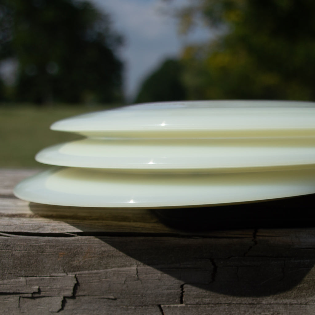 Three Realm - NXT Superglow distance drivers from Gateway Disc Sports, light-colored and stacked on a weathered wooden surface outdoors with trees and a cloudy sky in the background.