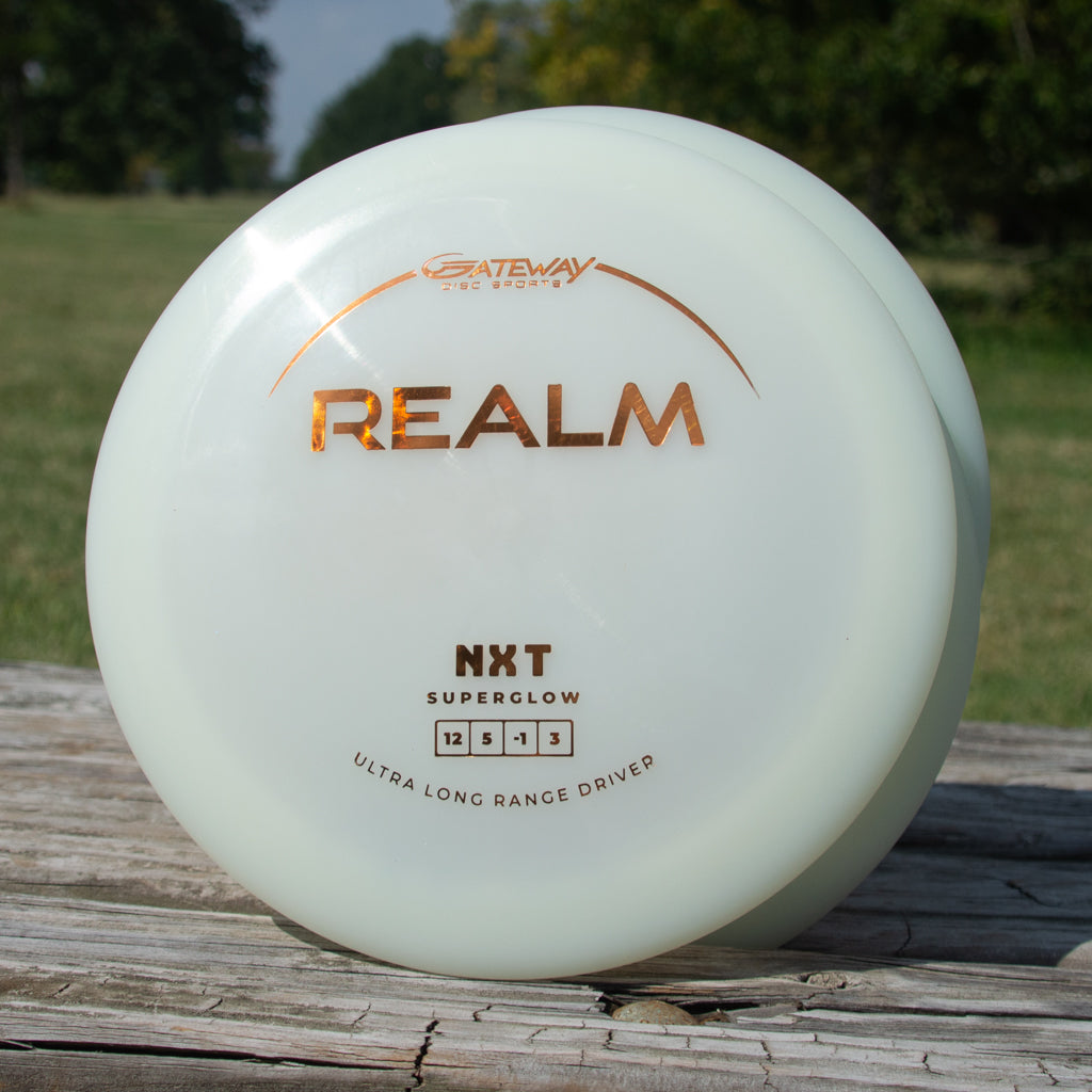 A white Realm - NXT Superglow max distance driver by Gateway Disc Sports is displayed upright on a wooden outdoor surface with trees and grass in the background.