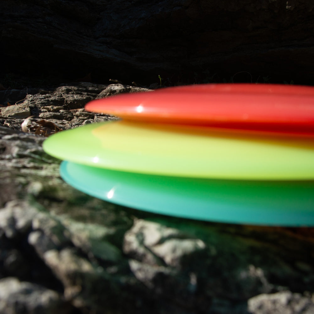 Three stacked Realm - Diamond frisbees in red, yellow, and green—ideal for power throwers or max distance—rest on a rocky surface with a blurred background.