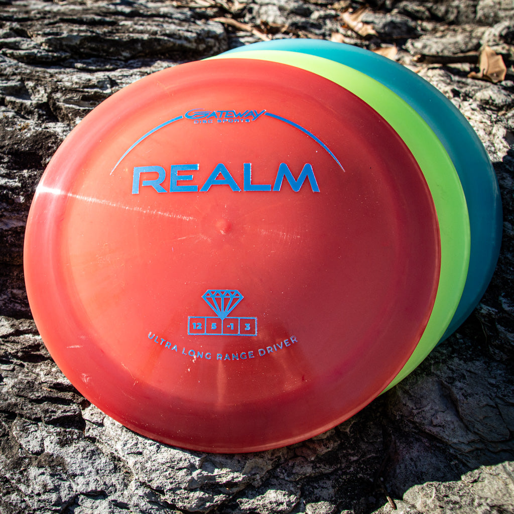 A red Realm - Diamond max distance driver rests atop two other discs, all placed on rough tree bark.