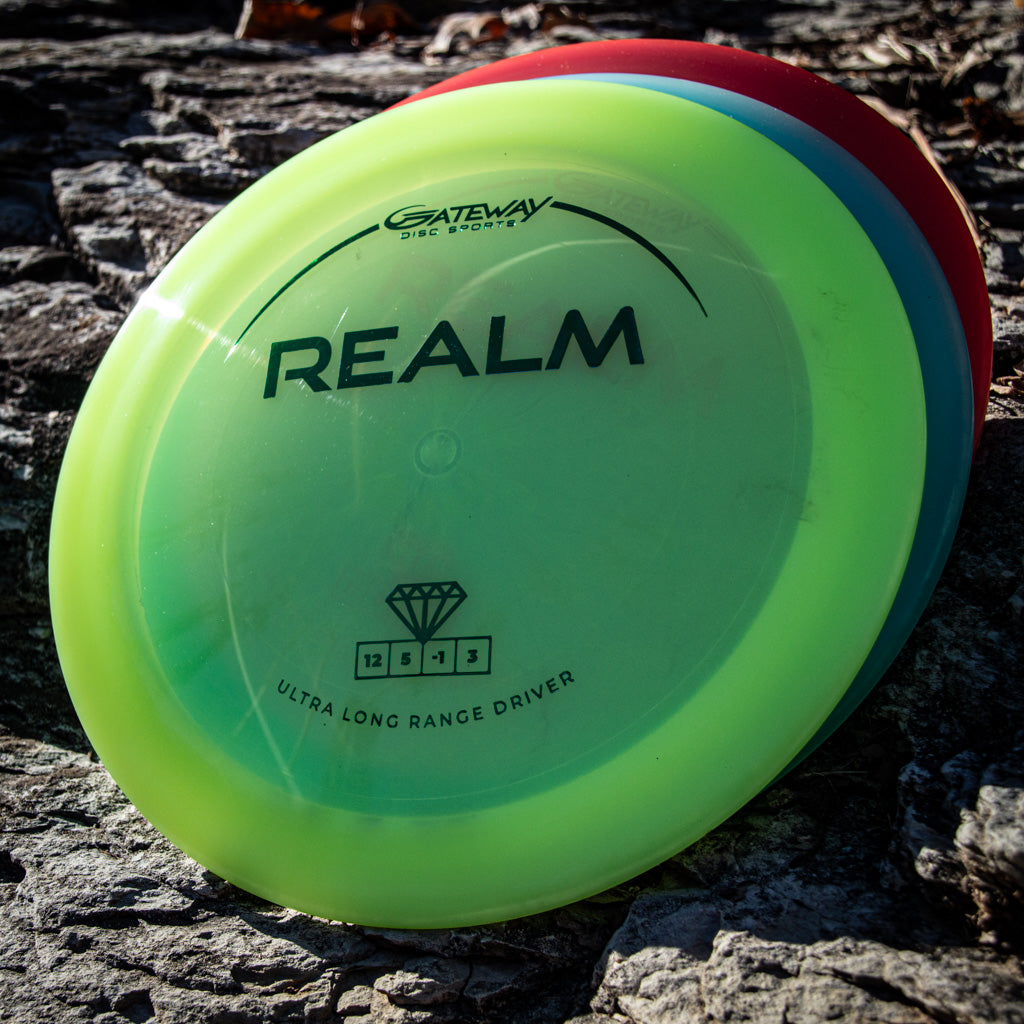 Three disc golf drivers rest on sunlit rocks, the front labeled "Realm - Diamond" by Gateway Disc Sports—a trusted distance driver ideal for power throwers.