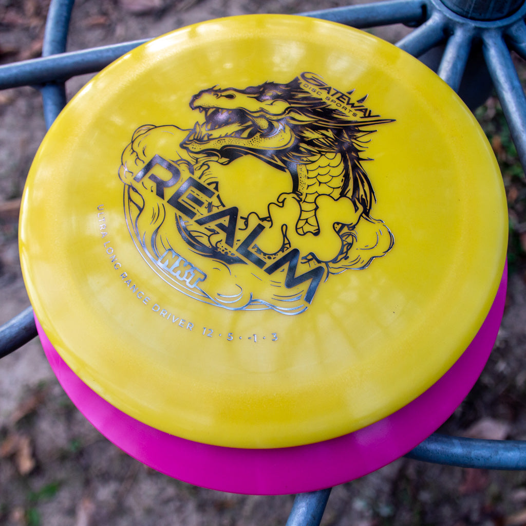 A yellow Realm NXT - Dragon distance driver with a dragon design and "REALM" text, made from durable plastic, rests atop a pink disc on an outdoor disc golf basket.