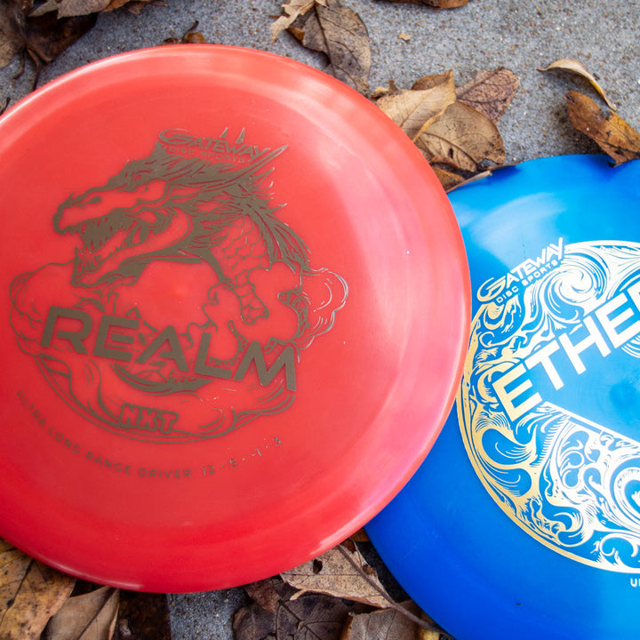 A red Realm NXT - Dragon distance driver from Gateway Disc Sports, made of durable NXT plastic and featuring a dragon design, lies among dry leaves on the ground.