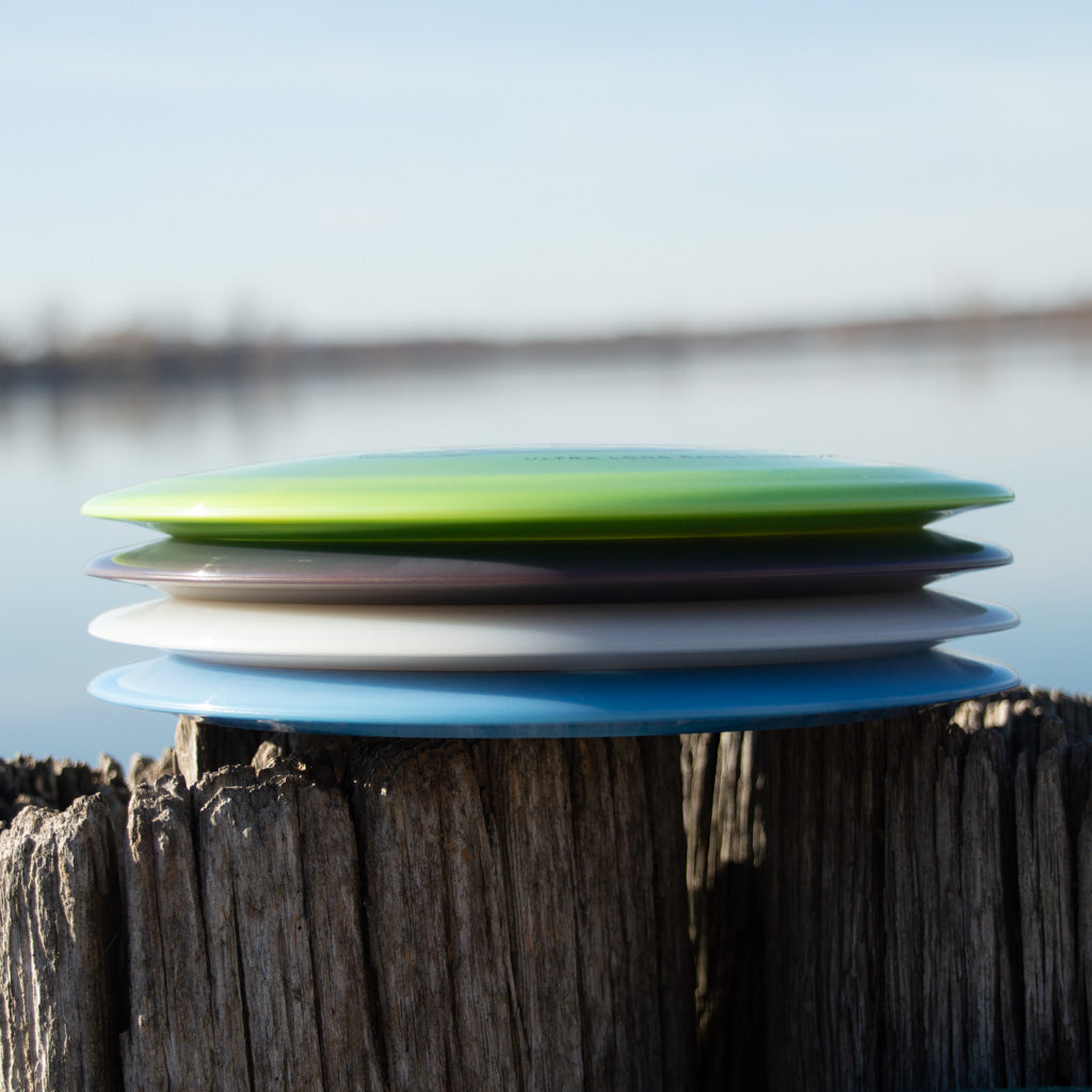 A stack of five colorful discs, including the Realm NXT - Dragon distance driver, sits on a weathered wooden post by a calm lake with a blurred shoreline in the background.