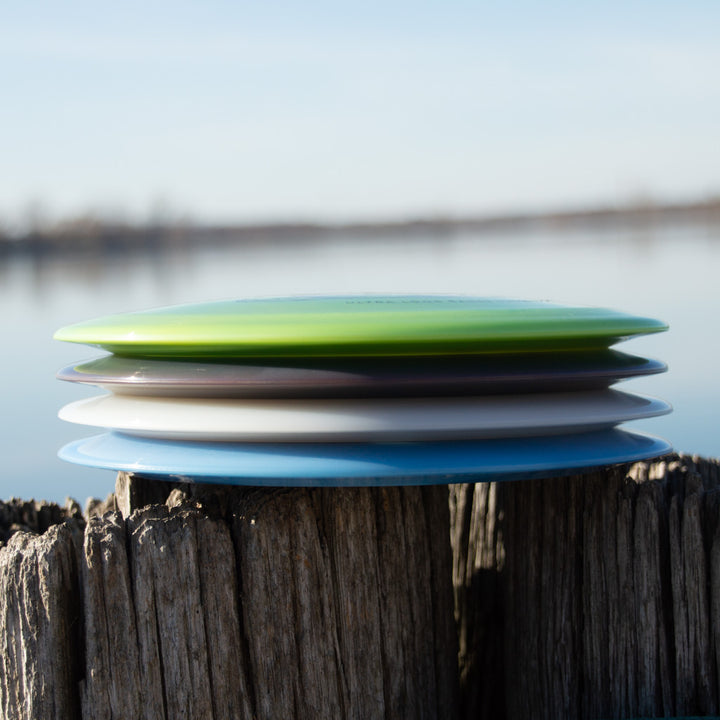 A stack of five colorful discs, including the Realm NXT - Dragon distance driver, sits on a weathered wooden post by a calm lake with a blurred shoreline in the background.