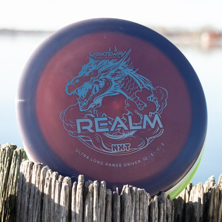 A Realm NXT - Dragon distance driver by Gateway Disc Sports, maroon with a silver dragon and "REALM NXT" design in durable NXT plastic, sits on a weathered wooden post near the water.