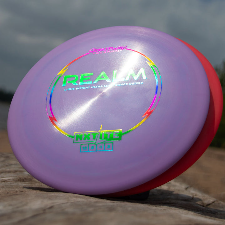 The Realm - NXT LITE, a purple distance driver with holographic "REALM" text, and a red disc stand upright on a log outdoors, highlighting the quality of Gateway Disc Sports.