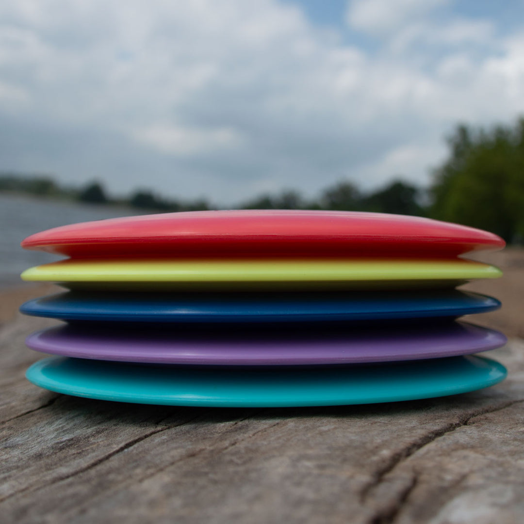 A stack of six colorful Realm - NXT LITE discs from Gateway Disc Sports sits outdoors on wood, with trees and water in the background—ideal for anyone seeking a reliable max distance driver.