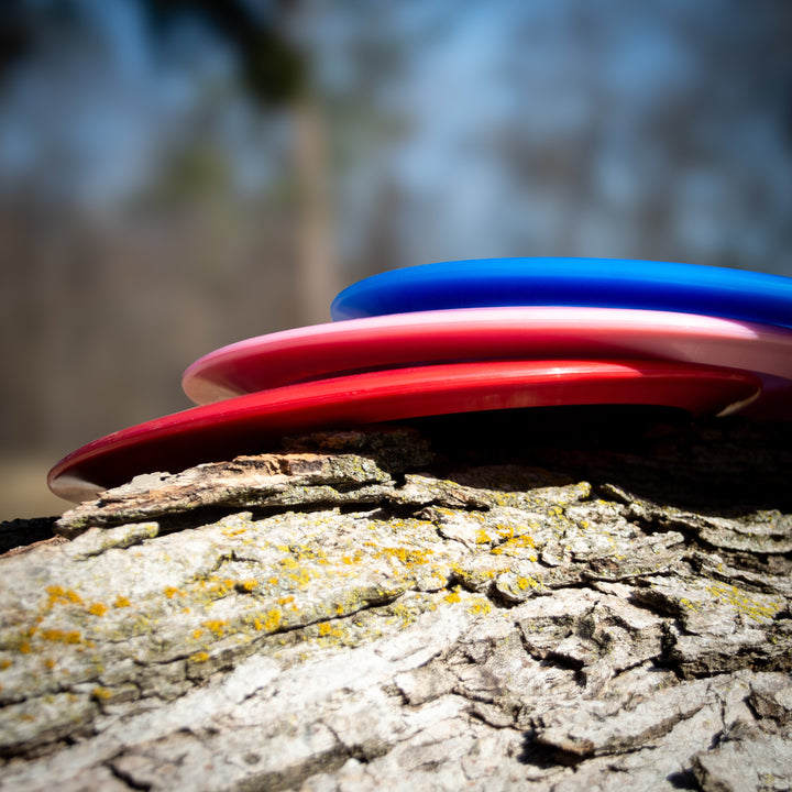 A blue Realm - Platinum distance driver disc and two red discs are stacked on a tree trunk outdoors, with a blurred background.