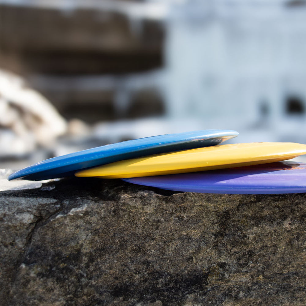 Three vibrant Realm - Suregrip Ultra Lightweight disc golf discs—blue, yellow, and purple—are stacked on a large rock with a blurred outdoor backdrop, perfect for power throwers seeking the ultimate distance driver.