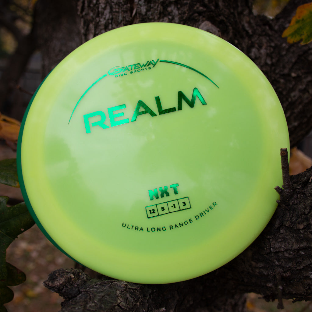 A yellow-green Gateway Disc Sports "Realm - NXT" distance driver (flight ratings: 12, 5, -3, 3), made from durable plastic, rests on a tree branch—perfect for power throwers.