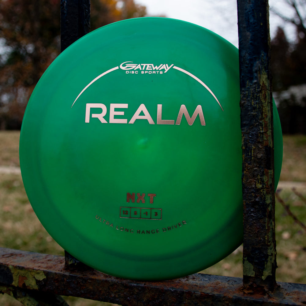 A green Realm - NXT distance driver by Gateway Disc Sports, made from durable plastic for power throwers, is shown outdoors against a metal fence with grass and trees in the background.