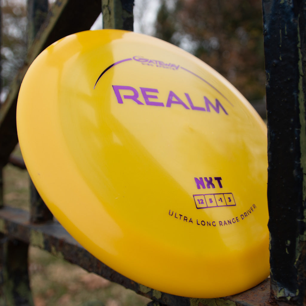 A yellow Realm - NXT distance driver made from durable plastic rests against a metal fence outdoors.