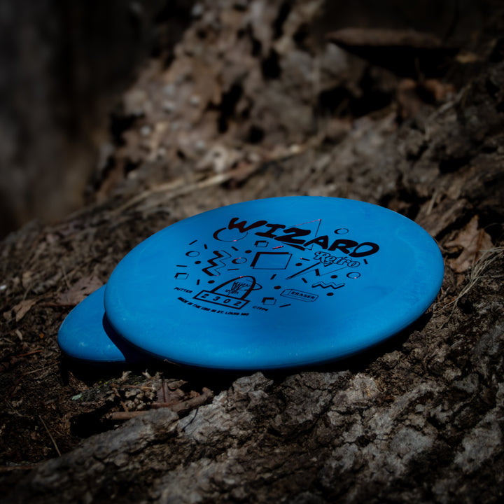 Two blue Retro Wizard - Eraser discs sit on rough tree bark among dry leaves, ideal for use as putters or approach discs.
