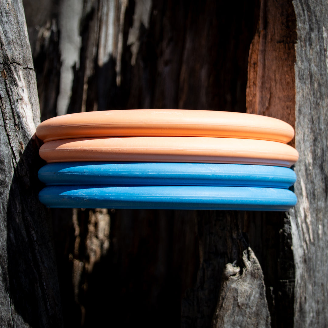 Four Retro Wizard - Eraser discs—two orange, two blue—from the Retro series are stacked on a rough wood surface, illuminated by bright sunlight.