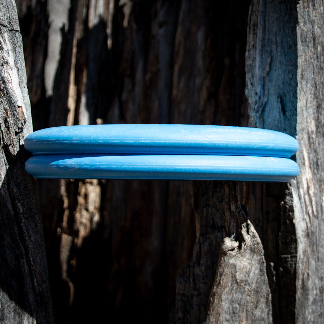 A blue Retro Wizard - Eraser disc is wedged vertically between two pieces of weathered tree bark in sunlight.