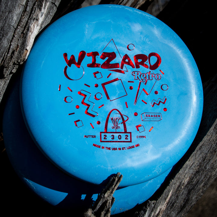 A blue Retro Wizard - Eraser disc golf putter with red geometric designs and text, partially stacked on another putter, rests on weathered wood.