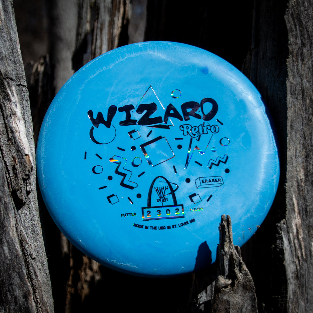 A Retro Wizard - Eraser disc golf putter in blue with black graphics stands upright against a weathered wooden surface outdoors, highlighting the classic charm of the Retro Wizard series.