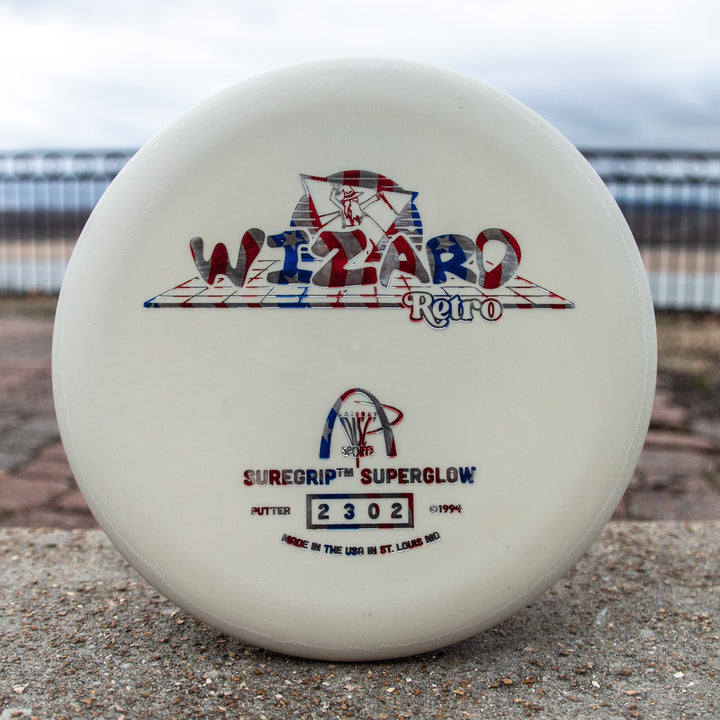 A Retro Wizard - SS Superglow disc golf putter in white features red, blue, and silver "Suregrip Superglow" and "2302" graphics with Gateway feel, resting outdoors on concrete.