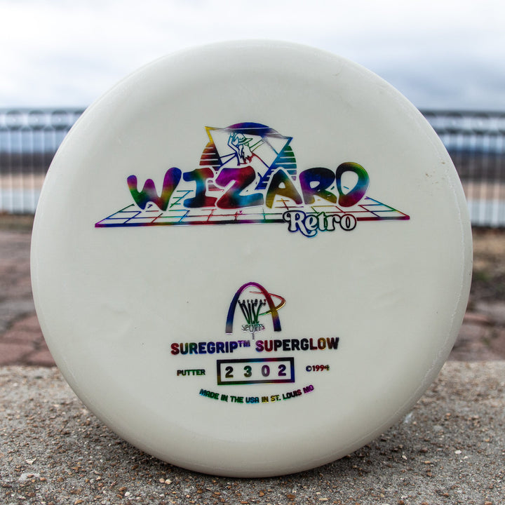 The Retro Wizard - SS Superglow is a white disc golf putter with colorful "Wizard Retro" text and logos, crafted with the Superglow formula for classic Gateway feel, plus "SureGrip SuperGlow" and serial number 2302 on the front.