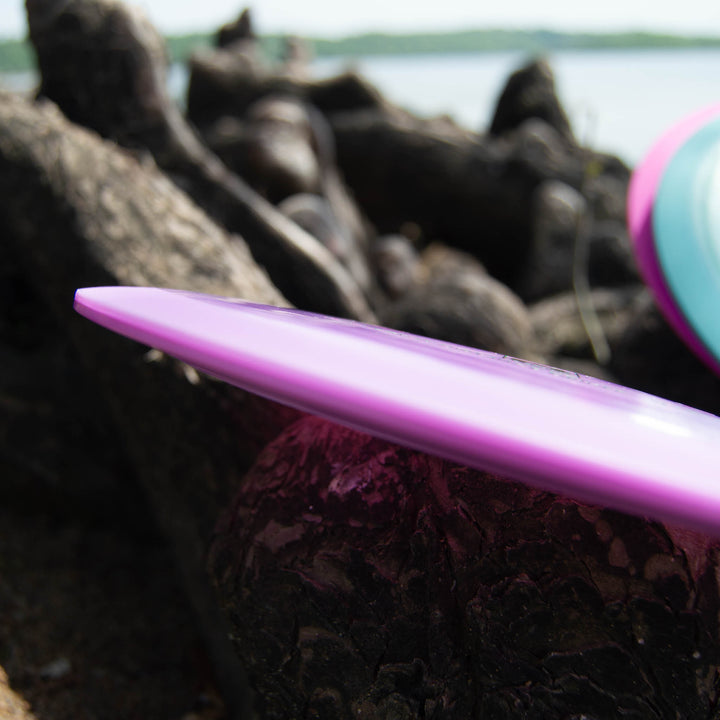 A close-up of the Samurai - Platinum, a purple distance driver frisbee crafted from eco-friendly plastic, resting on a textured outdoor surface with water and greenery in the background.