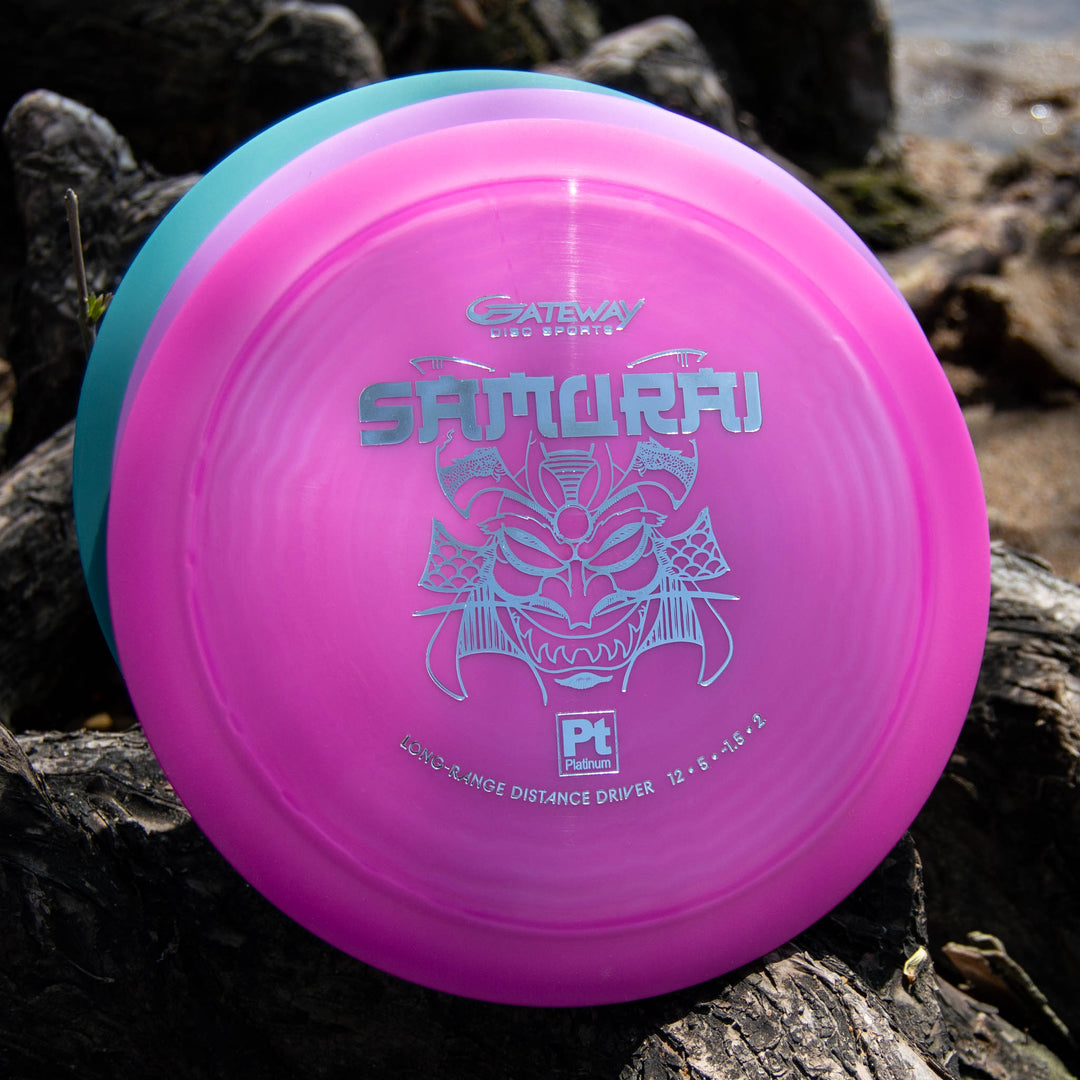 A pink Platinum Samurai distance driver by Gateway Disc Sports, made from eco-friendly plastic with a silver samurai helmet design, rests on textured tree roots outdoors.