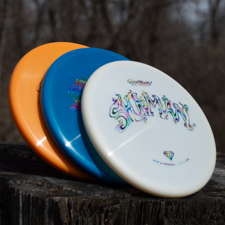 Three disc golf discs—orange, blue, and white—are stacked on a weathered wooden surface outdoors. The top white disc is the Shaman - Diamond, ready for its next flight down the fairway.