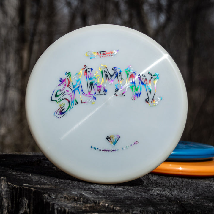A white Shaman - Diamond disc golf putter with vibrant, stylized text on its face stands upright on a tree stump outdoors—an ideal choice for fans of the Wizard series seeking precise, reliable performance.
