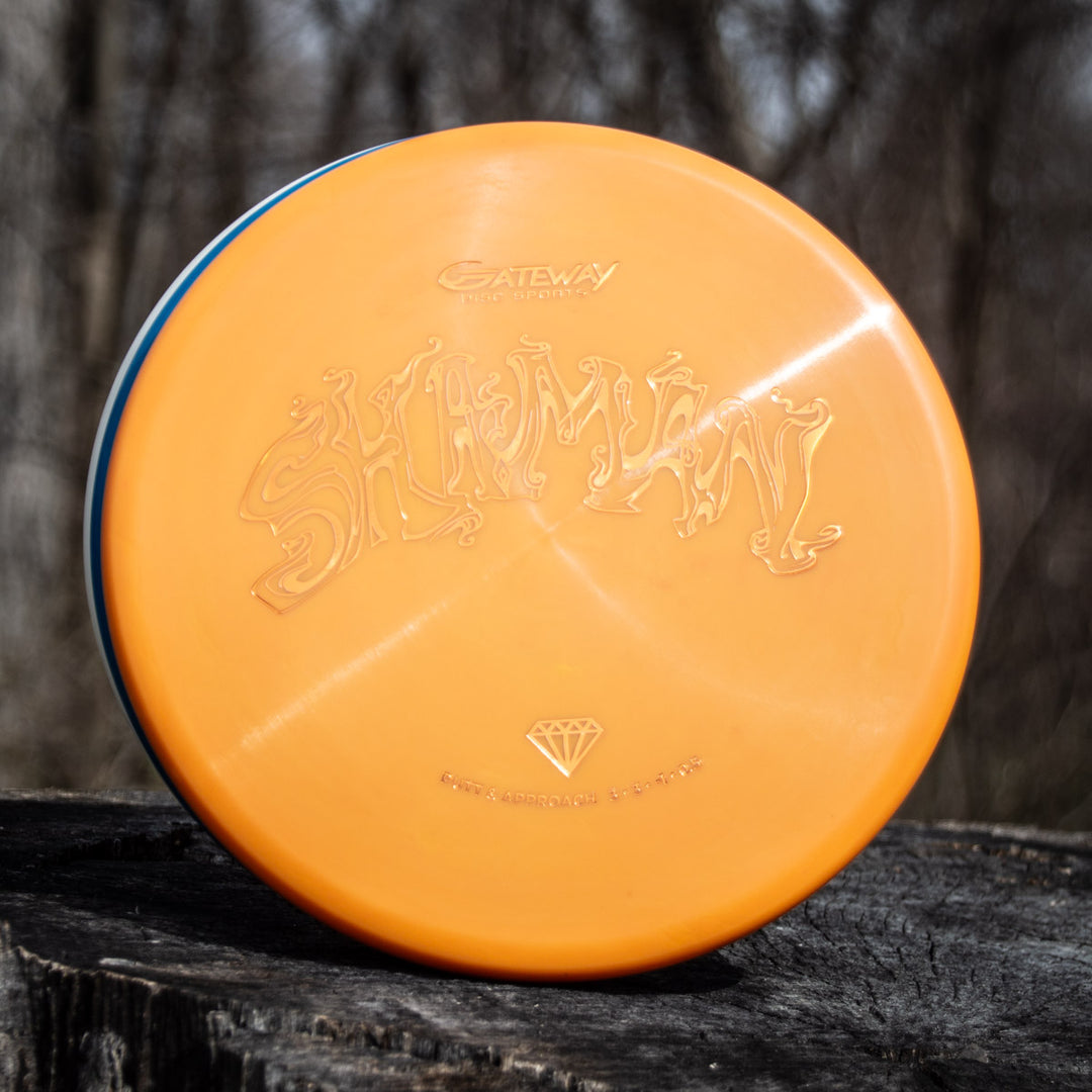 A Shaman - Diamond disc golf disc in orange stands upright on a tree stump, set against a blurred forest background, highlighting its distinctive style.