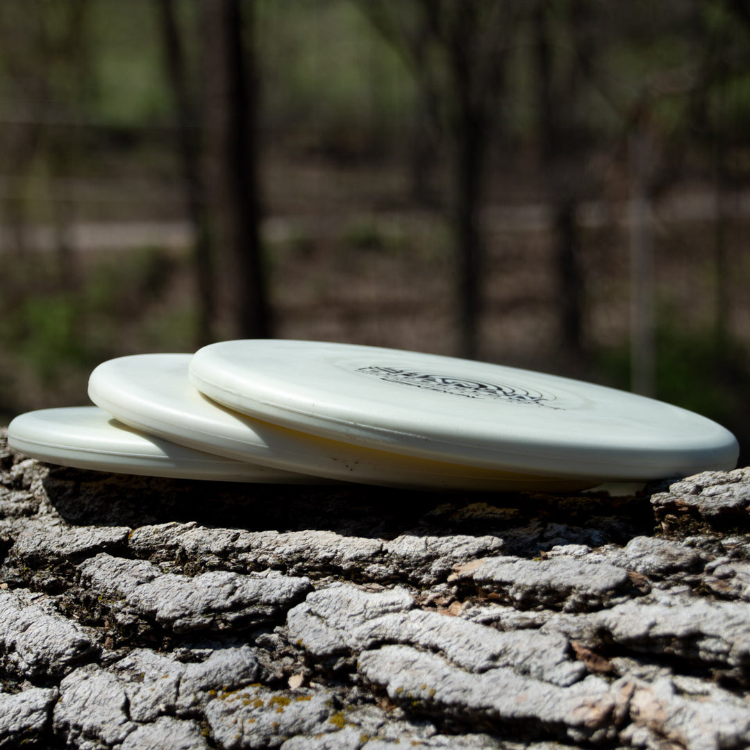 Three Shaman - SS - Suregrip Superglow putt and approach discs stacked on a textured tree trunk with a blurred forest background.