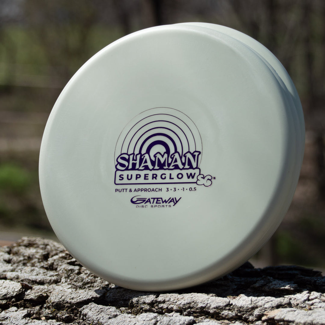 A white "Shaman - SS - Suregrip Superglow" putt and approach disc from Gateway Disc Sports, made with glow plastic, sits on a tree trunk outdoors.