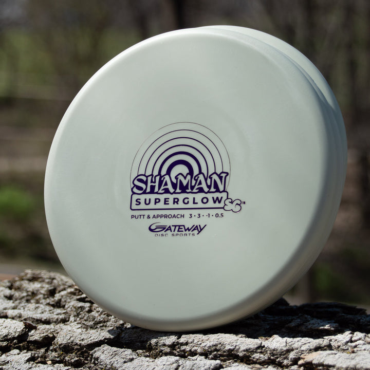 A white "Shaman - SS - Suregrip Superglow" putt and approach disc from Gateway Disc Sports, made with glow plastic, sits on a tree trunk outdoors.
