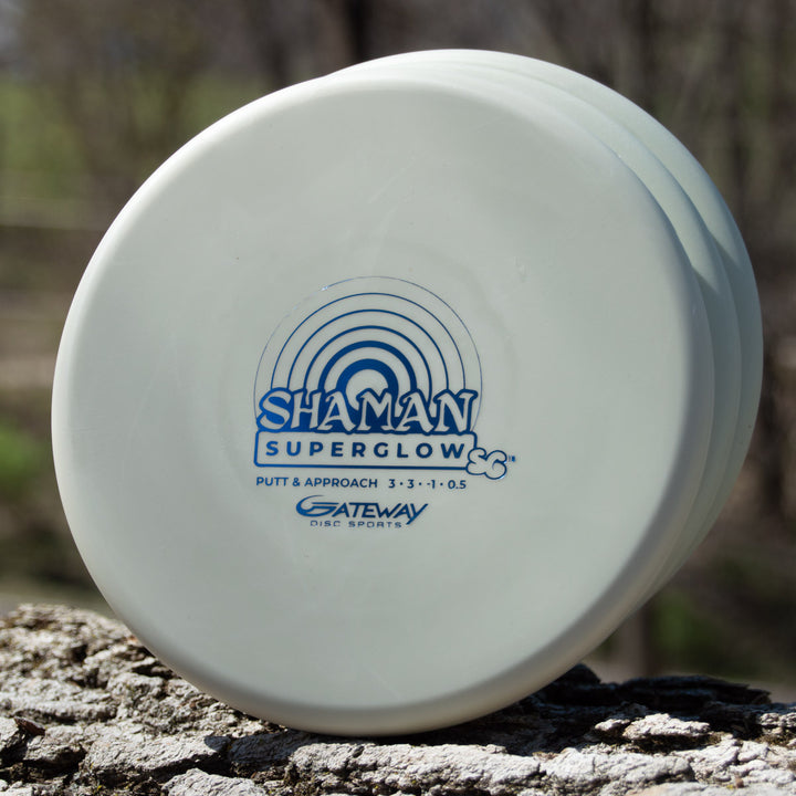 A white Shaman - SS - Suregrip Superglow putt and approach disc rests on a tree trunk, showing its blue logo and flight numbers on the durable glow plastic.