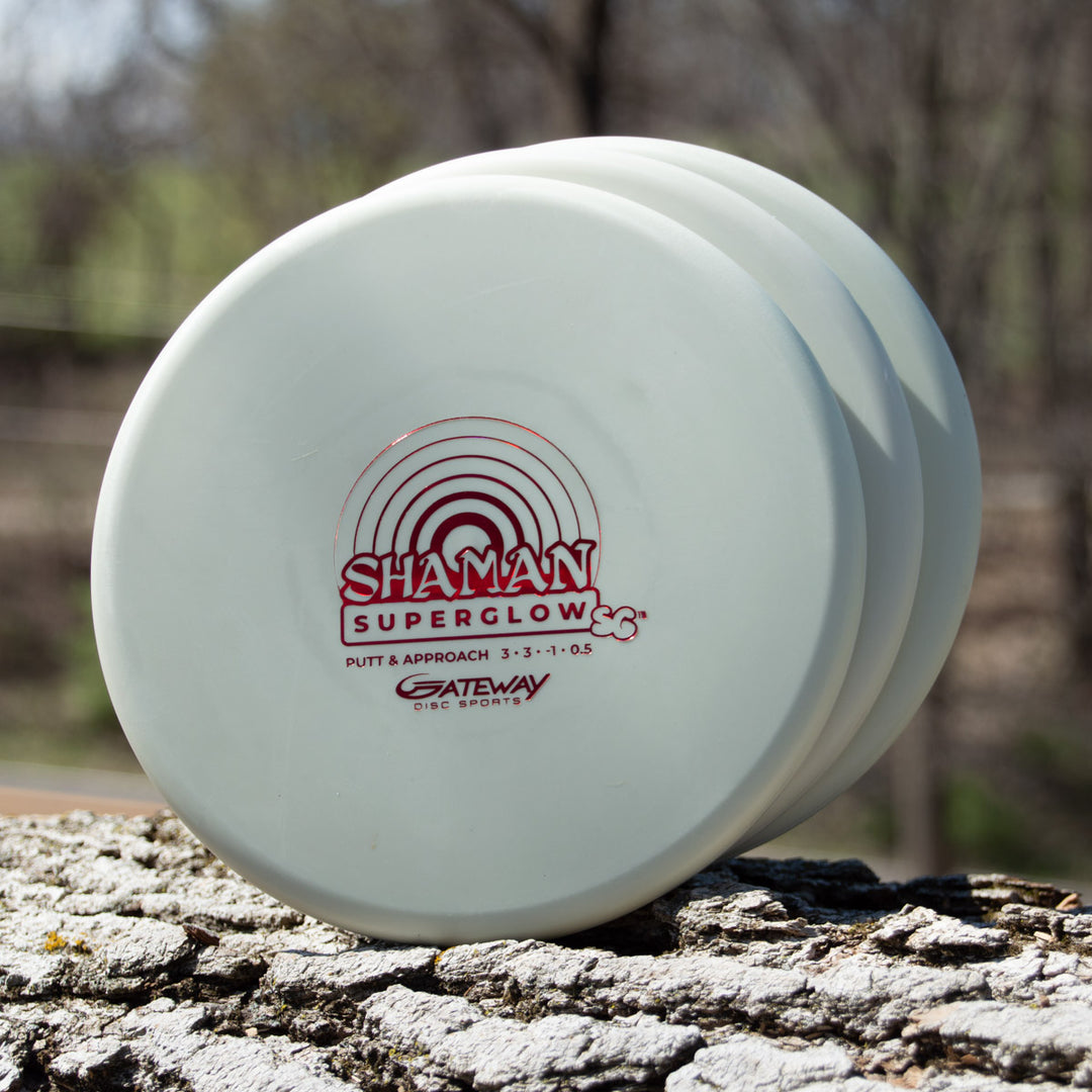 Three white Shaman - SS - Suregrip Superglow putt and approach discs made of glow plastic are stacked vertically on a tree trunk outdoors.