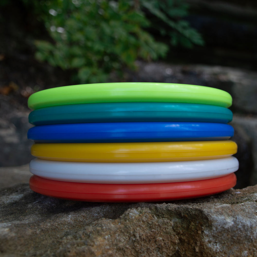 A stack of seven Shaman - NXT plastic frisbees in green, light blue, dark blue, yellow, white, orange, and red—with flat noses—rests on an outdoor stone surface.
