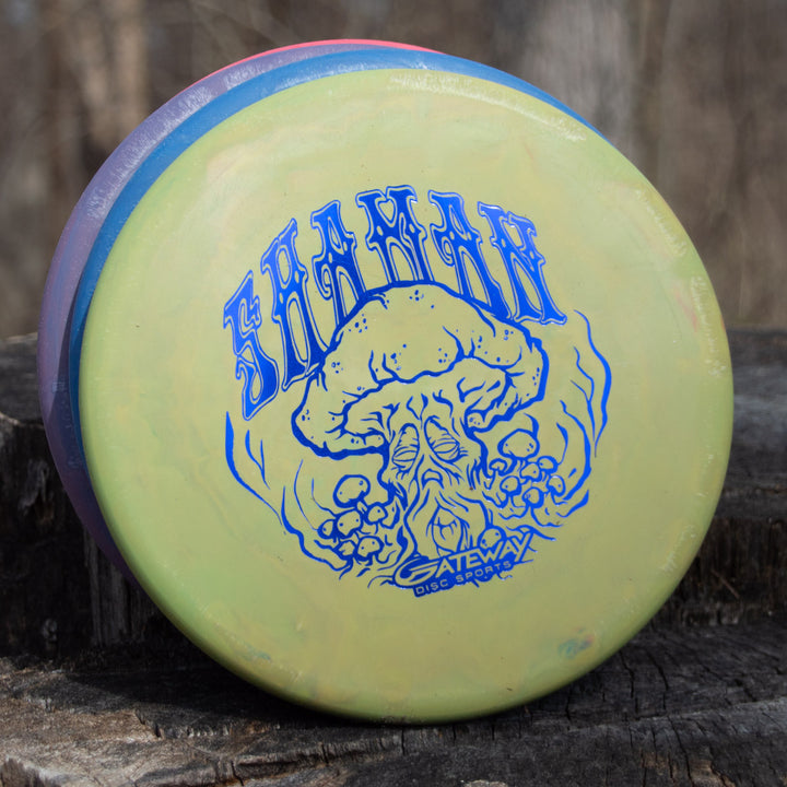 A yellow Shaman - SSS - Mushroom disc golf putter by Gateway Disc Sports, featuring a blue skull and mushroom Special Edition Stamp, sits on a wooden surface outdoors.