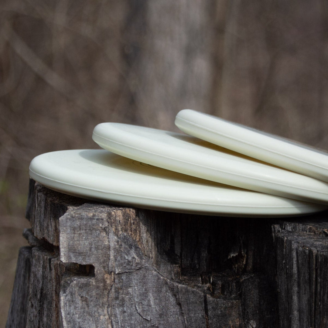 Three Shaman - SS - Suregrip Superglow putt and approach discs, crafted from glow plastic, are stacked on a tree stump outdoors with a blurred forest background.