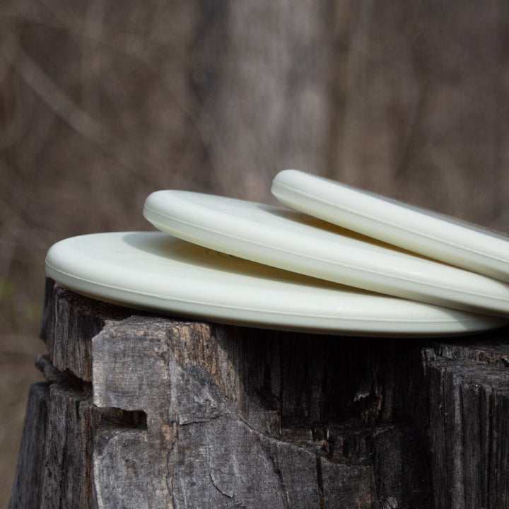Three Shaman - SS - Suregrip Superglow putt and approach discs, crafted from glow plastic, are stacked on a tree stump outdoors with a blurred forest background.